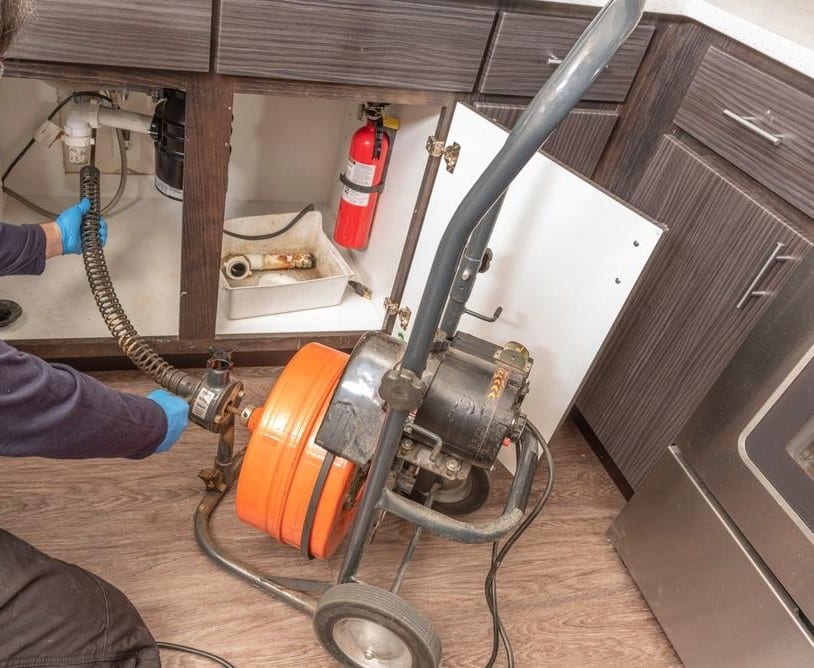 Technician feeding a drum auger cable into the drain beneath a kitchen sink