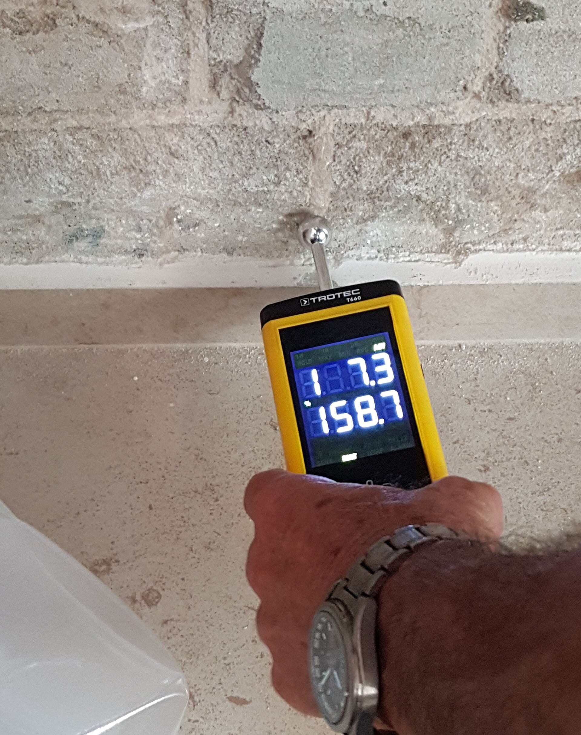 Moisture meter held against a wall displaying a live moisture reading